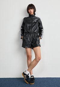 Chándal atlético de cuero negro con rayas blancas, chaqueta con cremallera y pantalones cortos a juego. Combinado con zapatillas negras con suela de goma.