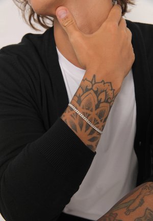 Silver chain bracelet worn on a tattooed forearm with a black cardigan and white t-shirt; intricate tattoo design visible.