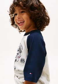 Χαμογελαστό νεαρό παιδί με σγουρά μαλλιά, που φοράει ένα λευκό και ναυτικό μπλουζάκι ράγκλαν με σχέδιο αθλητή του τένις καρτούν και αριθμό 85.