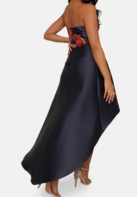 Nainen käyttää olkaimetonta kukallista tummaa laivastonsinistä satiinista korkeavyötäröistä helmatyylistä mekkoa, jossa on koristellut beiget korkokengät, käsi lepää alaselällä.