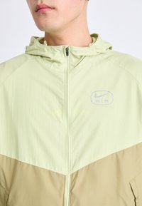 Lichtgroene en tan jas met een ritssluiting, capuchon en "AIR" logo. De stof lijkt lichtgewicht met een gladde textuur.