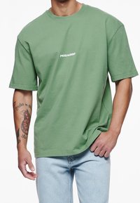 Muž v volném zeleném tričku s nápisem "PEGADOR" uprostřed hrudi a světle modrých džínech, viditelné tetování na levém paži.