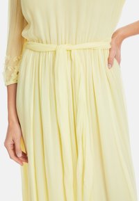 Helles gelbes Kleid mit tailliertem Schnitt, Gürtelakzent, plissiertem Rock und durchsichtigen Ärmeln, die mit kleinen Blumenmotiven verziert sind.