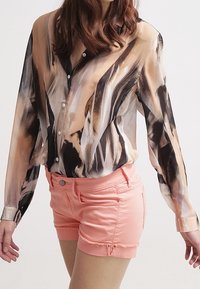 Camicia trasparente a maniche lunghe con motivi astratti nei toni beige, marrone e nero, abbinata a pantaloni corti in denim rosa aderenti con orli sfrangiati.
