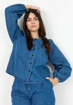 Frau mit langen dunklen Haaren, die ein blaues Jeans-Top mit Knöpfen und passende Hose trägt, posiert mit einer Hand am Kopf und der anderen in der Tasche.