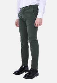 Chinos verde scuro in cotone, con vestibilità slim, tasche frontali, chiusura con un solo bottone, abbinati a scarpe eleganti nere lucide.