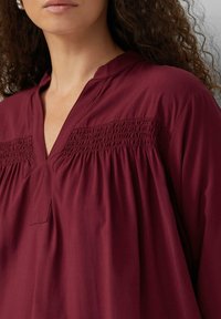 Bordeaux tuniek met een V-hals, voorzien van smockdetails op de schouders, lichte textuur en een losse, vloeiende pasvorm.