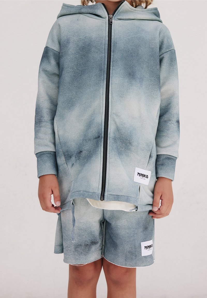 Enfant portant un sweat à capuche zippé tie-dye bleu-gris et un short assorti avec des patches logo Minikid blancs sur un fond uni.