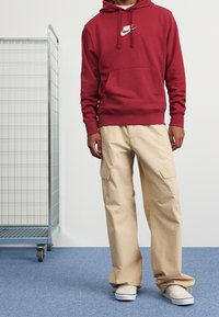 Mörkröd Nike-hoodie med framficka och logotyp, ihop med beiga cargobyxor med sidofickor och vita slip-on sneakers.
