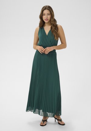 Jeune femme debout portant un haut sans manches vert profond à col en V et une jupe longue plissée assortie avec des sandales à talons noires.