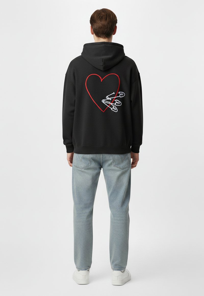 Felpa nera con cappuccio caratterizzata da una grande grafica a forma di cuore rosso sul retro, accentuata da spille di sicurezza bianche. Indossata con jeans azzurri chiari.