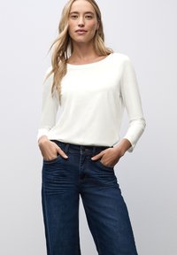 Langarm-weißes Oberteil mit Rundhalsausschnitt, kombiniert mit dunkelblauen, weit geschnittenen Jeans mit Fronttaschen und kontrastierenden Nähten.