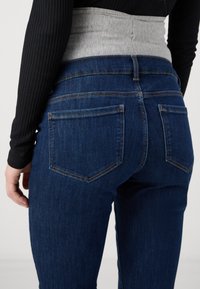 Vaqueros azul oscuro con bolsillos traseros, que presentan una cinturilla gris. La tela tiene una textura suave y un diseño clásico de pierna recta.