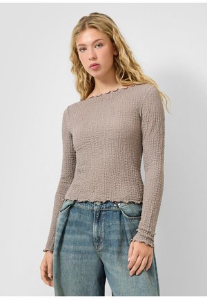 Młoda kobieta z długimi, falującymi blond włosami, ubrana w teksturowany beżowy top z długim rękawem i luźne niebieskie dżinsy, na jasnym, jednolitym tle.