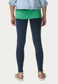 Leggings blu navy con una texture liscia, abbinati a una maglietta verde e a un leggero giubbotto di jeans, completati da sandali con cinturini color chiarissimo.