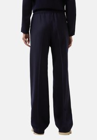 Persoon in een donkerblauwe wijde broek en bijpassend topje, staand met ontspannende armen tegen een effen lichte achtergrond.