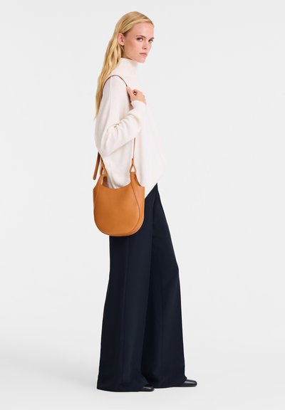 Tan leather crossbody bag with a curved shape and a single shoulder strap, paired with a cream knit sweater and navy wide-leg pants.
