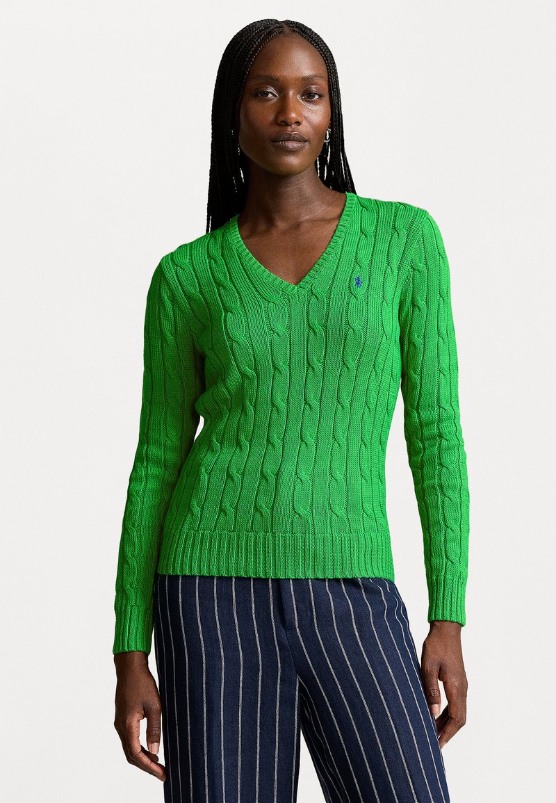 Pull en laine vert avec un col en V et des finitions côtes. Il a des manches longues et un petit logo sur la poitrine. Associé à un pantalon à rayures.