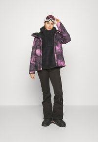 Skijacke mit lila Blumenmuster, schwarzem Fleece-Futter, kombiniert mit schwarzen Hosen und Schneestiefeln. Weiße Skibrille auf dem Kopf des Modells.