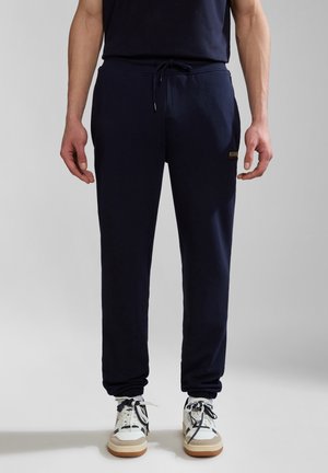 Mann trägt marineblaue Jogginghosen und weiße Turnschuhe, steht vor einem schlichten hellgrauen Hintergrund.