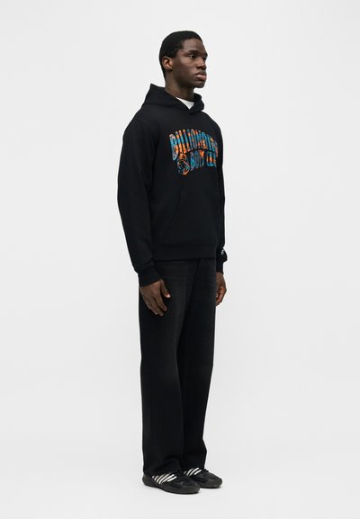 Άνδρας που στέκεται με μαύρη φόρμα με κουκούλα, με πολύχρωμο λογότυπο "Billionaire Boys Club", μαύρο παντελόνι και μαύρα αθλητικά παπούτσια με λευκές ρίγες σε γκρι φόντο.