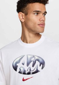 T-shirt en coton blanc avec un grand graphique "Air" en argent brillant et un petit emblème Nike en swoosh rouge en bas à droite.