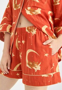 Röd pyjamaset med sydda guldfärger i en seriestil av sovande katter och månar, texturerat tyg och knappstängning på shorts.