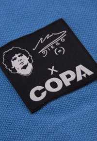Blauwe stof met een zwart vlak waarop in witte letters "COPA" staat, samen met een portret van Diego Maradona en zijn handtekening erboven.