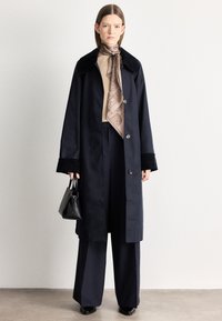 Μακρύ ναυτικό παλτό με μαύρο γιακά, συνδυασμένο με μπεζ τοπ και φουλάρι με σχέδια. Το μοντέλο κρατάει μια μικρή μαύρη τσάντα με γυαλιστερό φινίρισμα.