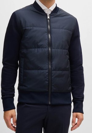 Hombre que lleva una chaqueta bomber acolchada azul marino con cremallera sobre una camisa blanca con cuello, mostrado desde el cuello hasta las caderas sobre un fondo liso.