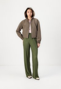 Chaqueta con capucha de color verde oliva, recortada, con mangas largas y cremallera frontal. Pantalones verdes plisados de cintura alta. Tela suave, fondo neutro.
