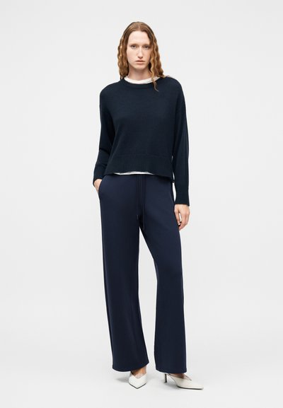 Γυναίκα με κυματιστά μαλλιά που φοράει μπλε σκούρο πουλόβερ, παντελόνα στο ίδιο χρώμα, λευκό υποκάμισο και λευκά παπούτσια με τακούνι, στέκεται μπροστά σε απλό φόντο.