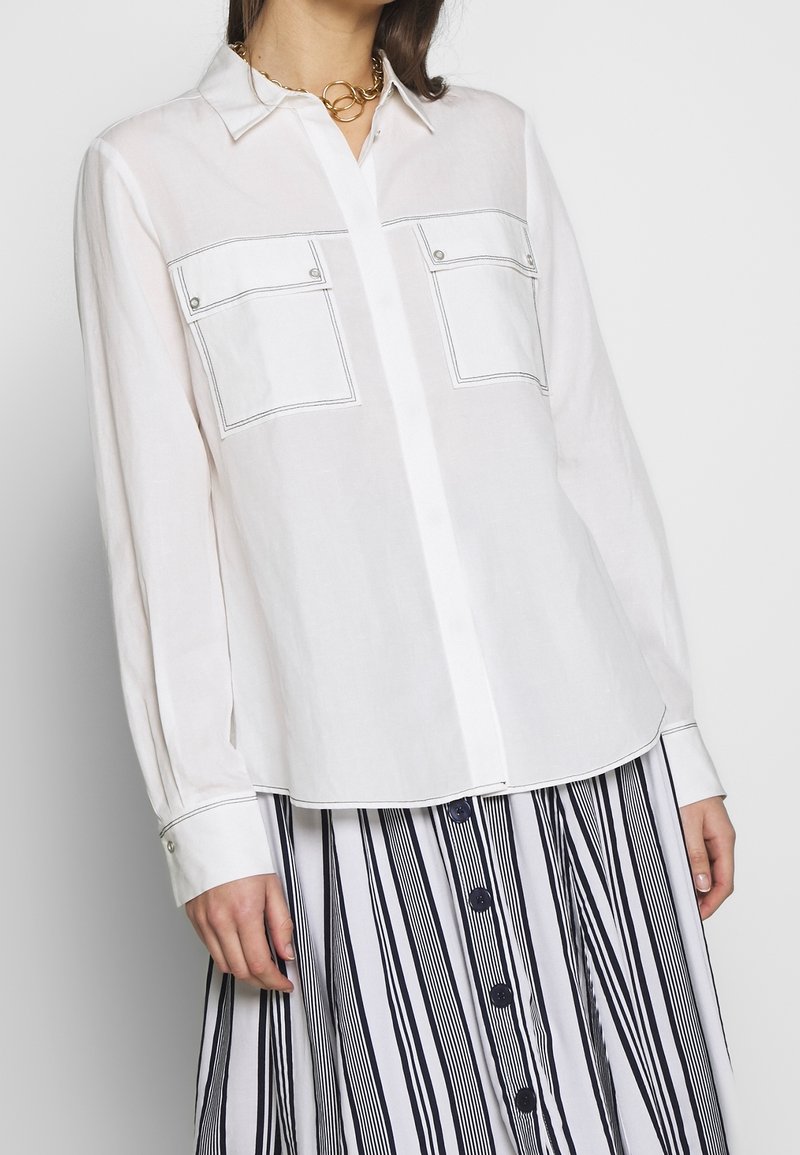 White button-up shirt with two chest pockets, contrast stitching, and long sleeves. Worn with a striped skirt featuring vertical lines.