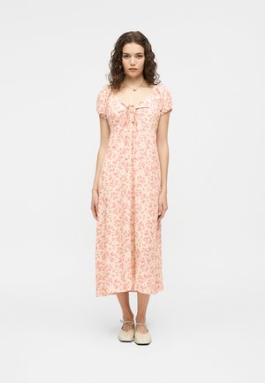 Νεαρή γυναίκα με σγουρά μαλλιά, φορώντας ένα ροζ ανοιχτό φόρεμα μέσης μήκους με φλοράλ σχέδιο και φουσκωτά μανίκια, καθώς και κρεμ επίπεδα παπούτσια, στέκεται μπροστά σε λευκό φόντο.