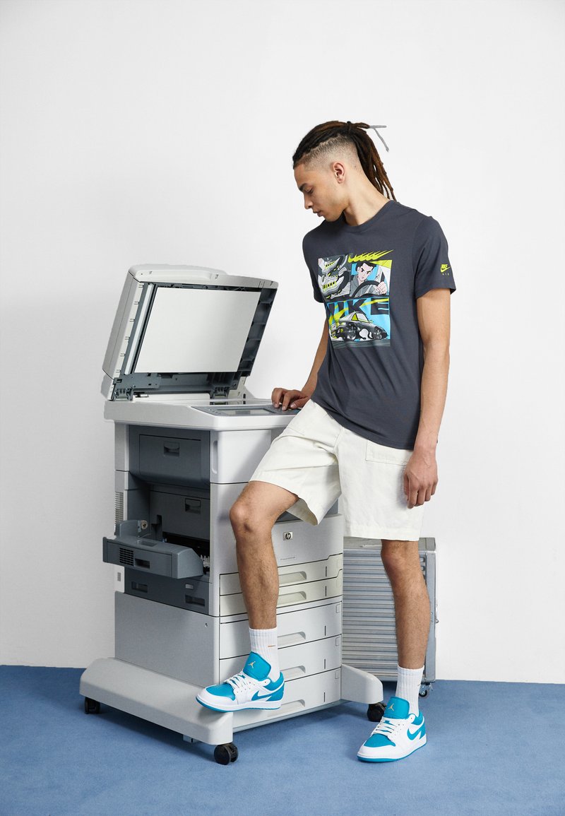 Graues T-Shirt mit einem bunten Grafikdesign, weiße Shorts und blaue Turnschuhe. Die Personen interagieren in der Nähe eines grauen Multifunktionsdruckers.