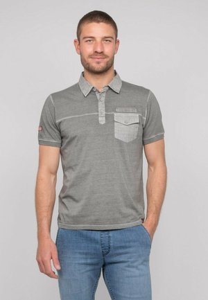 Homme souriant, portant un polo gris à manches courtes avec une poche poitrine et un jean bleu délavé, debout devant un fond clair uni.