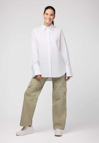 Camicia bianca oversize con bottoni e polsini allungati, abbinata a pantaloni cargo beige chiaro con tasche laterali, indossata con sneakers bianche.