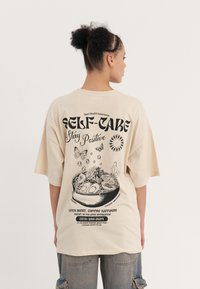 Beige oversized t-shirt featuring a black graphic print on the back, showcasing a bowl, plants, and butterflies, with text elements.