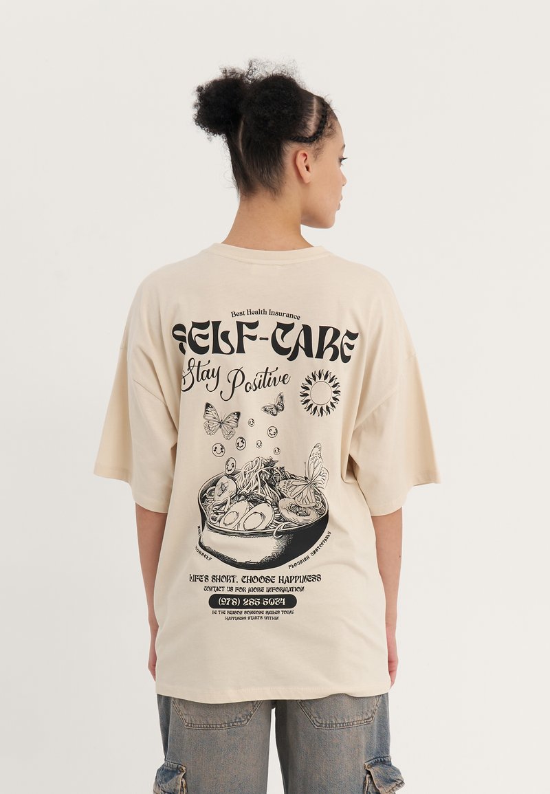 Beige Oversized-T-Shirt mit einem schwarzen Grafikdruck auf der Rückseite, der eine Schüssel, Pflanzen und Schmetterlinge zeigt, mit Textelementen.