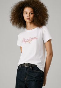 T-shirt en coton blanc avec le logo "Pepe Jeans" en rose, à manches courtes et col rond, associé à un jean en denim foncé et une ceinture noire.
