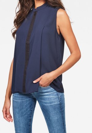 Kvinde iført navyblå ærmeløs bluse med sort lodret kant, kombineret med lyseblå jeans, hånd i lommen, mod en hvid baggrund.