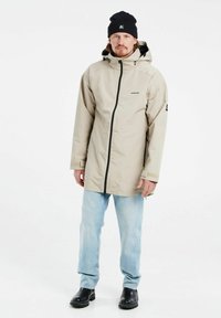 Giacca impermeabile beige con cappuccio, zip frontale e dettaglio logo. Abbinata a jeans azzurri chiari e stivaletti neri. Texture liscia.
