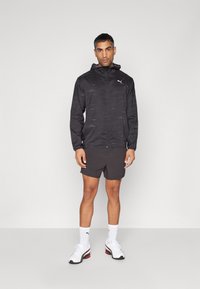 Svart hettejakke med strukturert mønster, grå shorts, hvite treningssko og hvite sokker. Har hette og PUMA-logo på brystet.