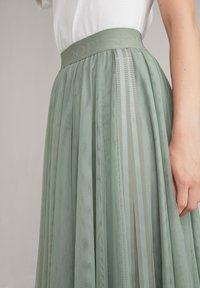 Pitkä, laskostettu hame vaaleanvihreästä tiulista, leveällä vyötärönauhalla, jossa on kohokuvioitu "JOOP!" ja sivuraitoja kontrastiväreissä.