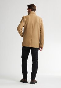 Cappotto di lana color sabbia con una vestibilità sartoriale, colletto alto e linee pulite, abbinato a jeans neri e stivali marroni. Design minimalista e texture liscia.