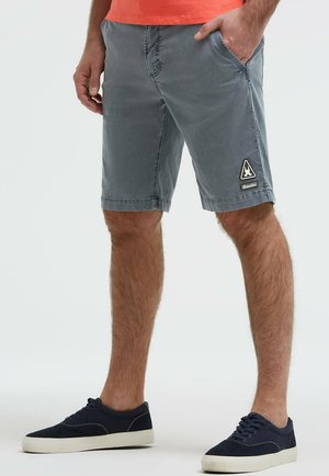 Mann trägt graue knielange Shorts mit Seitentaschen und schwarze Turnschuhe, steht vor einem schlichten Hintergrund.