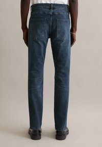 Jean en denim bleu foncé à coupe droite, avec deux poches arrière et un léger délavé. Porté avec des chaussures noires sur un fond neutre.