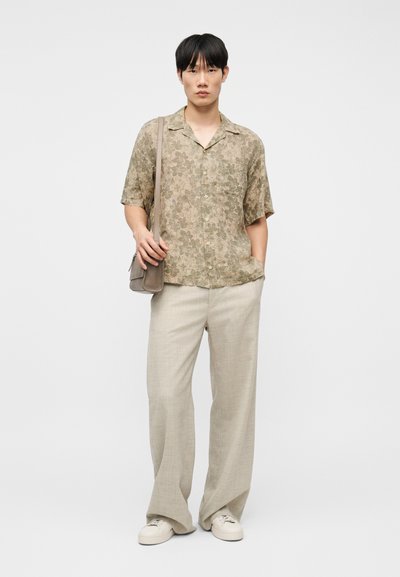 Νεαρός άνδρας που φοράει μπεζ φλοράλ πουκάμισο με κοντά μανίκια, φαρδιά ανοιχτόχρωμα καφέ παντελόνια, λευκά αθλητικά παπούτσια και κρατά μια ώχρα τσάντα ώμου, στέκεται μπροστά σε λευκό φόντο.