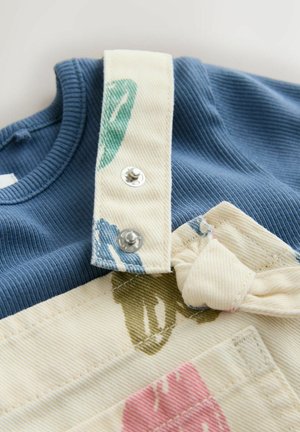 Blue ribbed shirt collar paired with cream overalls featuring green and pink floral patterns, secured by silver snaps and a tied waistband.