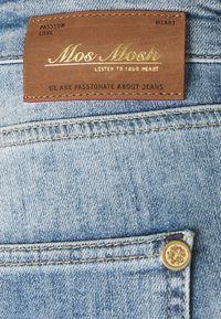 Světle modré džíny z denimu s hnědou koženou etiketou s raženým zlatým textem. Obsahují detail zlatého knoflíku na pasu.